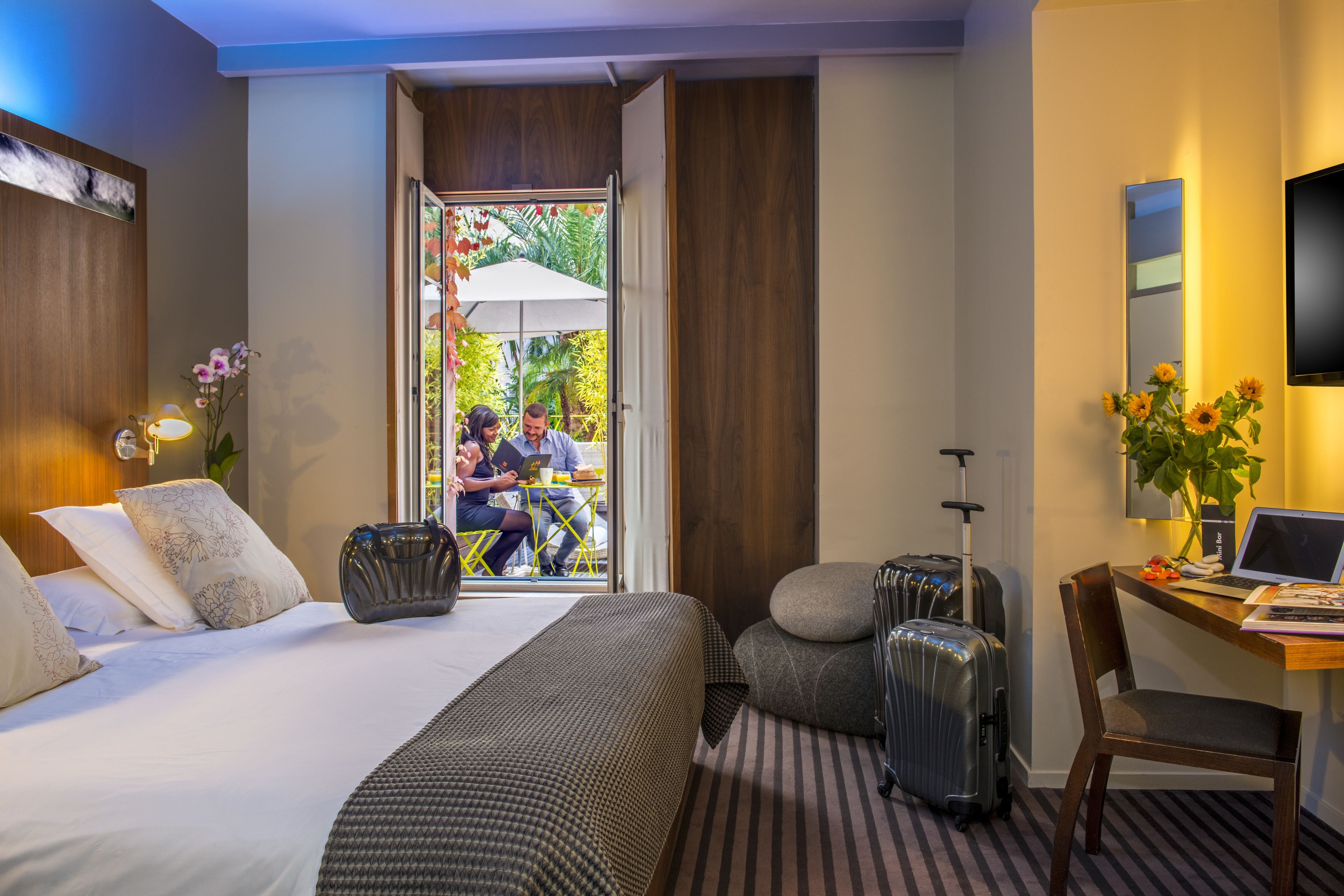 A hotel room with a bed, a desk with a laptop, a chair, and two suitcases.