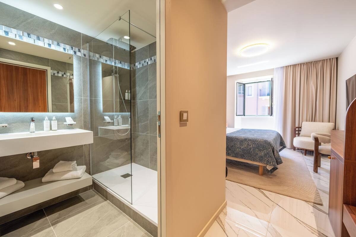 A modern bathroom with a glass shower, a vanity with toiletries, and a bed in the adjacent room.