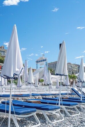 Zona de playa con hileras de sombrillas blancas y sillas reclinables azules, cerca de una valla de piquetes blancos y un edificio con un letrero al fondo.