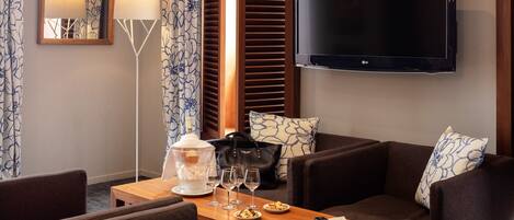 Habitación de hotel con televisión de pantalla plana, una mesita de madera con aperitivos y bebidas, un sofá con cojines estampados y una lámpara de pie.