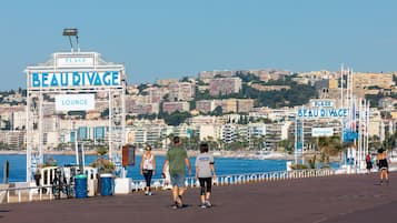 Um calçadão com placas de "BEAU RIVAGE" e "PLAGE", vista do horizonte urbano e uma praia ao fundo.
