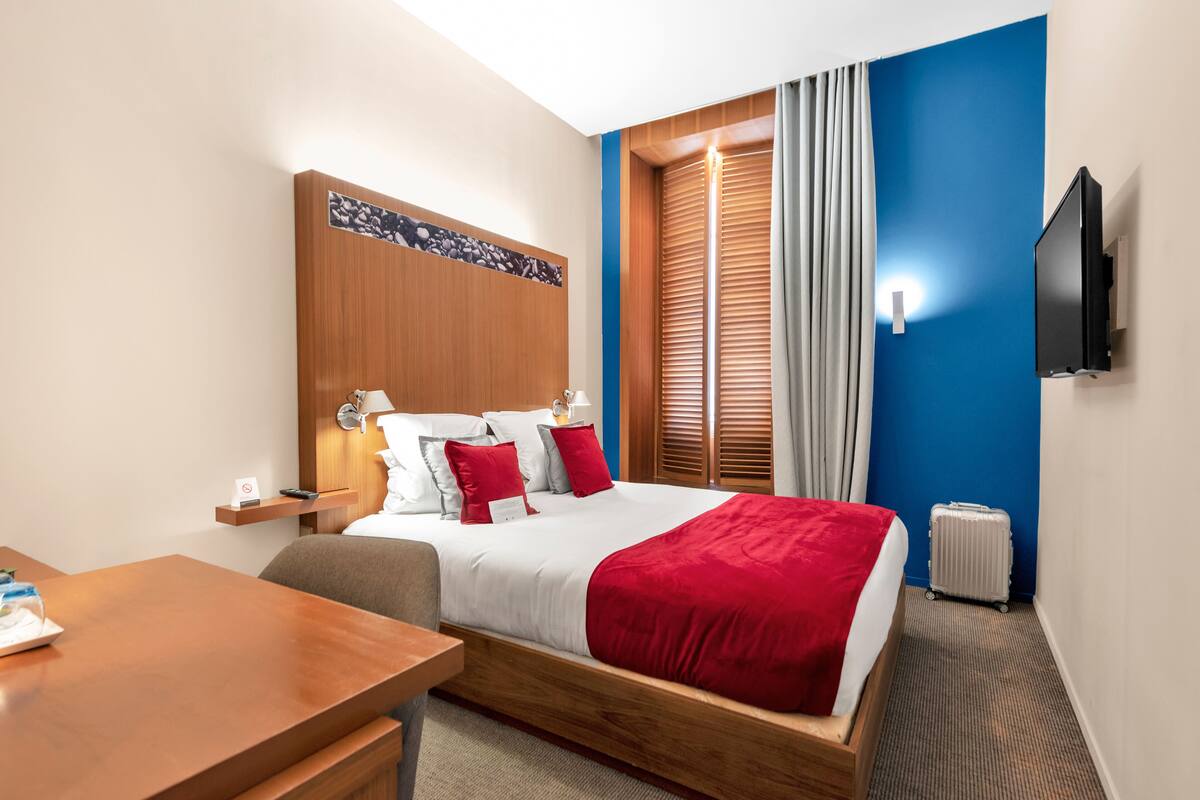 A hotel room with a bed, bedside table, lamp, TV, and a suitcase.