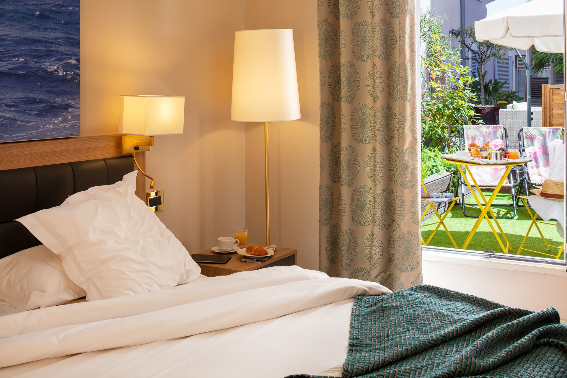 Una habitación de hotel con una cama, lámparas de noche, una mesita con una taza y un vaso, y vistas al exterior con una mesa y sillas.
