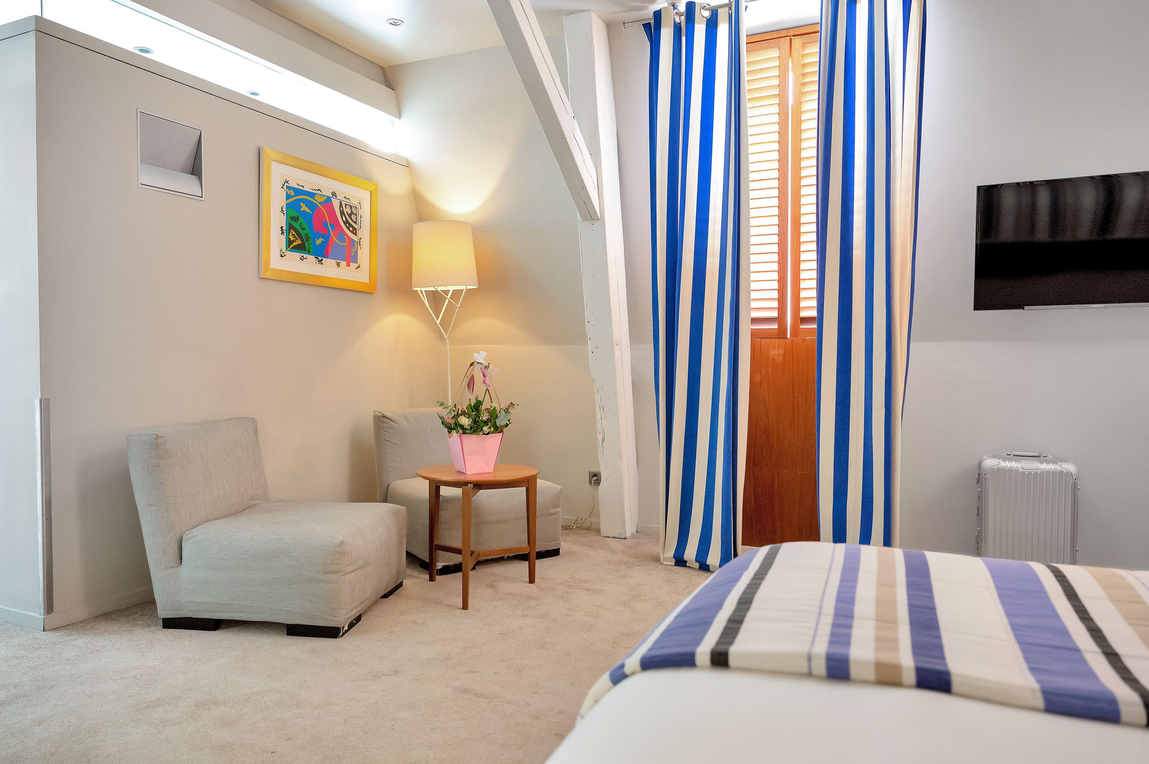 A hotel room with a bed, a chair, a small table with a plant, a painting on the wall, and a TV mounted on the wall.