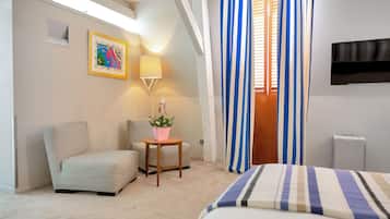 A hotel room with a bed, a chair, a small table with a plant, a painting on the wall, and a TV mounted on the wall.