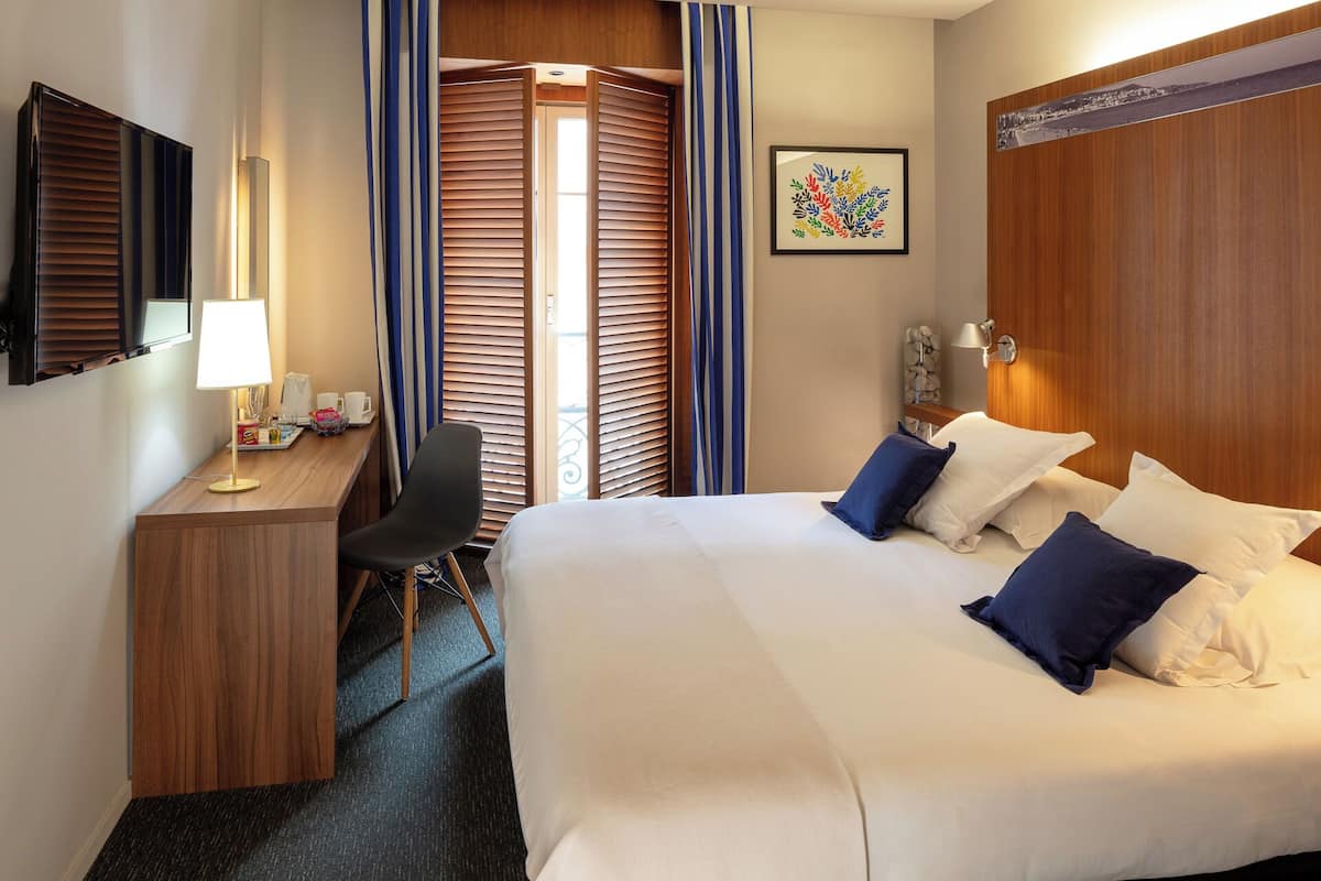 A hotel room with a bed, a desk with a lamp, a chair, and a window with blinds.