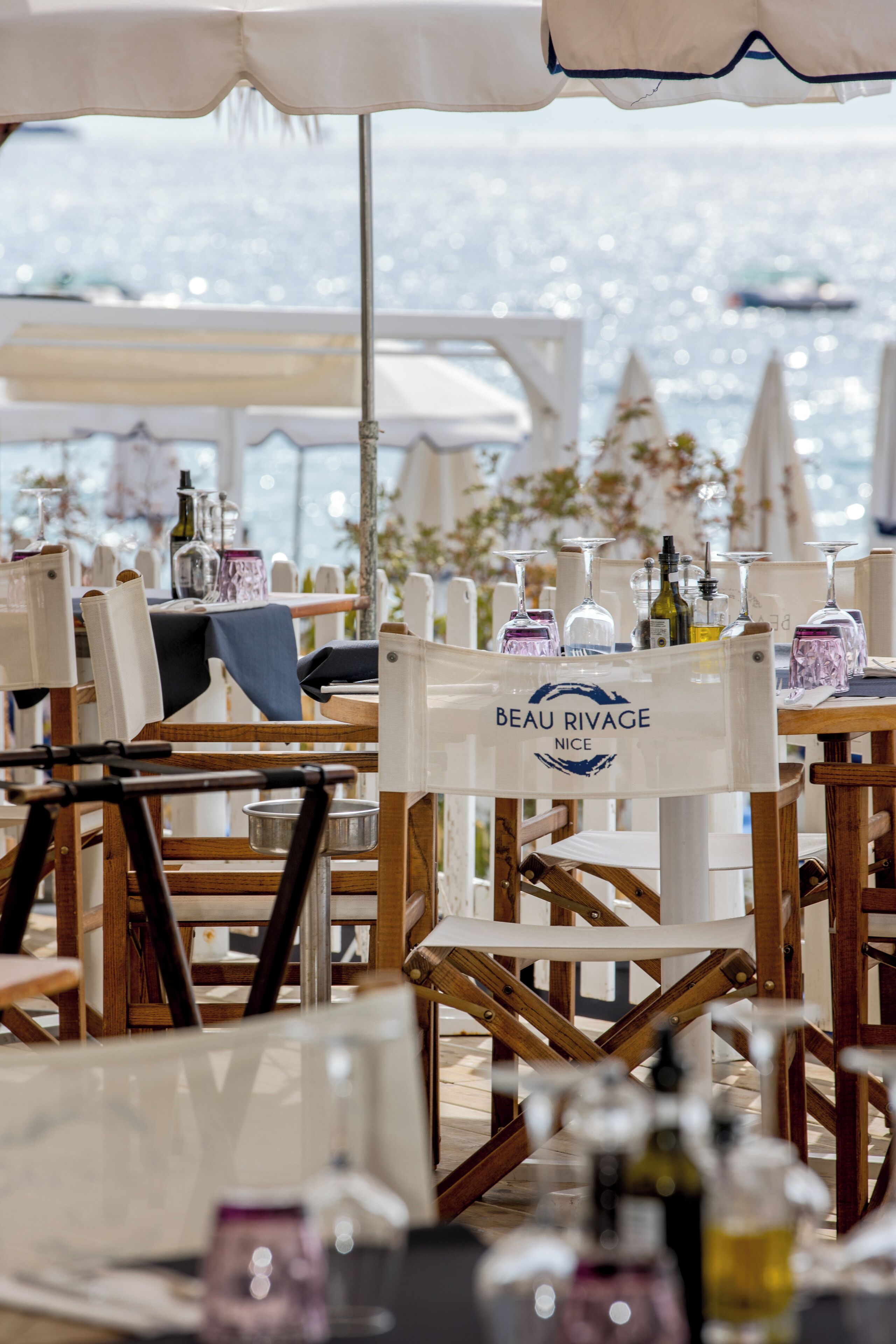 A dining area with wooden chairs and tables, some with umbrellas. A sign reads "BEAU RIVAGE NICE."