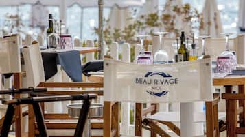 A dining area with wooden chairs and tables, some with umbrellas. A sign reads "BEAU RIVAGE NICE."