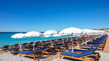 Een strand met rijen houten ligbedden en witte parasols, uitgestald op een kiezelstrand.
