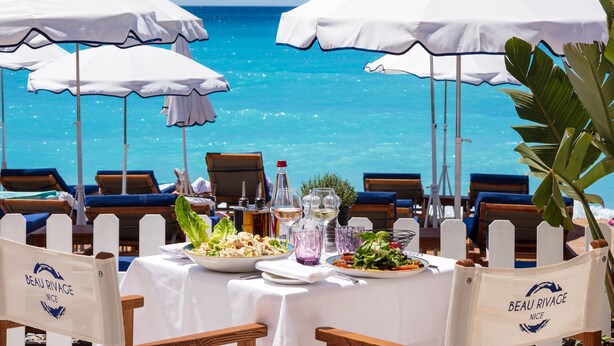 Una mesa para dos con vista al océano, rodeada de sombrillas blancas, y con la etiqueta 'Beau Rivage Nice'.