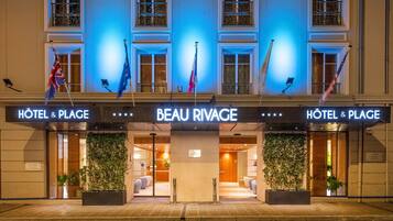 The exterior of the Beau Rivage hotel with a prominent entrance, two flags, and a sign indicating it is a hotel and plage.