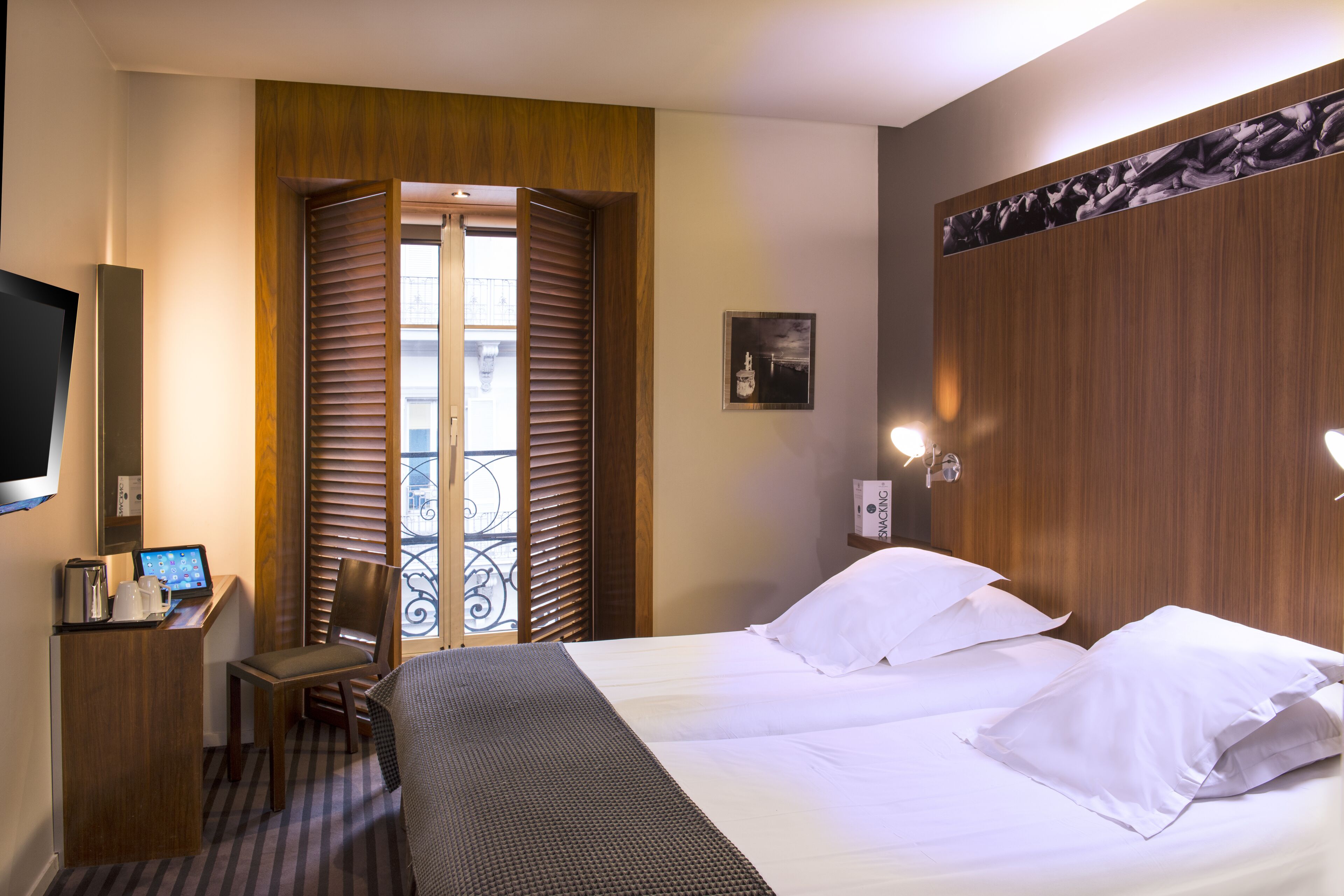 A hotel room with a bed, a desk with a computer, a chair, and a window with blinds.