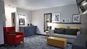 Une chambre d’hôtel moderne avec un fauteuil rouge, un canapé gris, une table basse en bois, un coin cuisine équipé d’un évier et de placards, et des œuvres d’art encadrées accrochées aux murs.