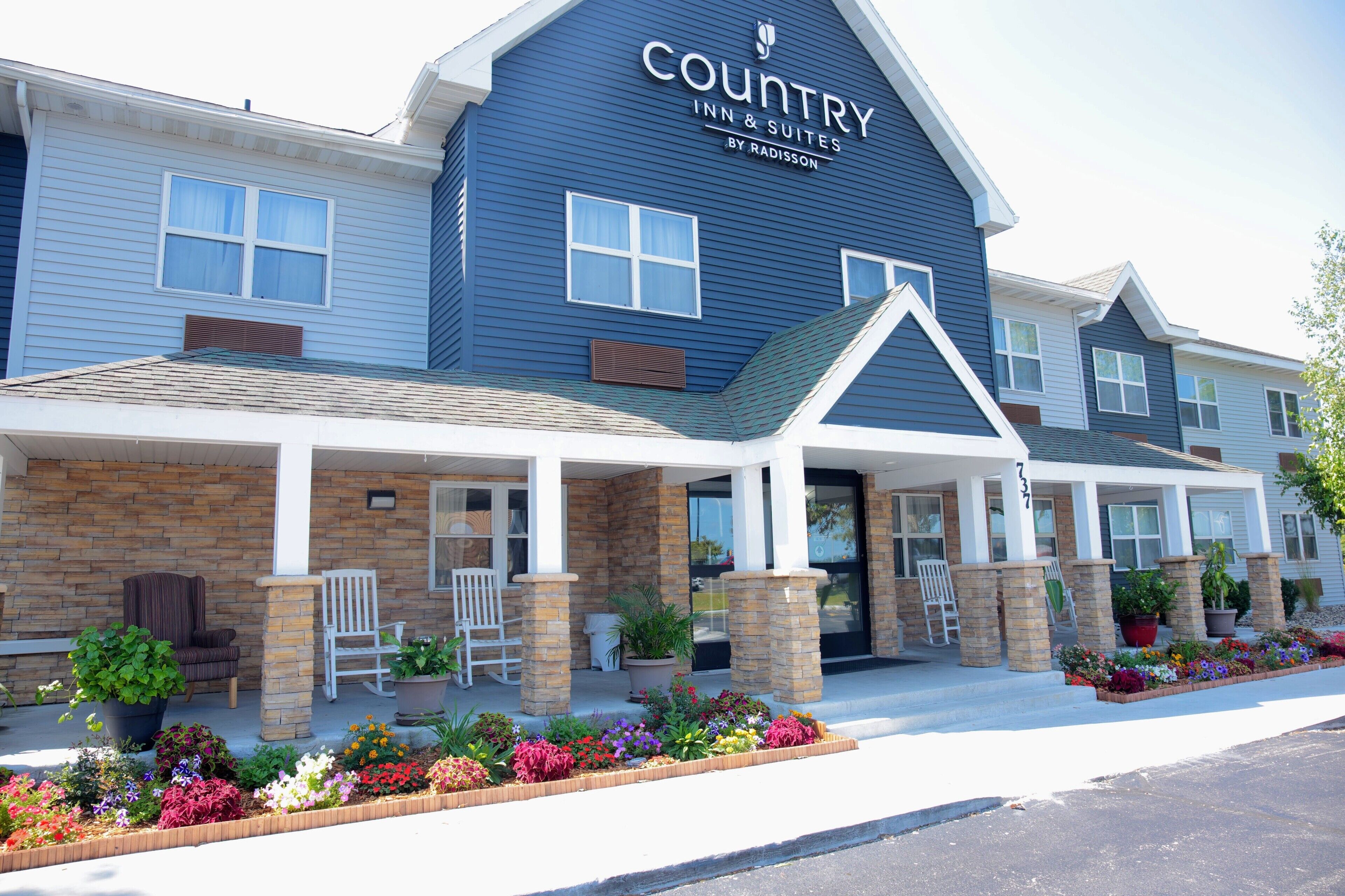 A Country Inn & Suites by Radisson with a covered front porch, outdoor seating, and a variety of potted plants and flowers.