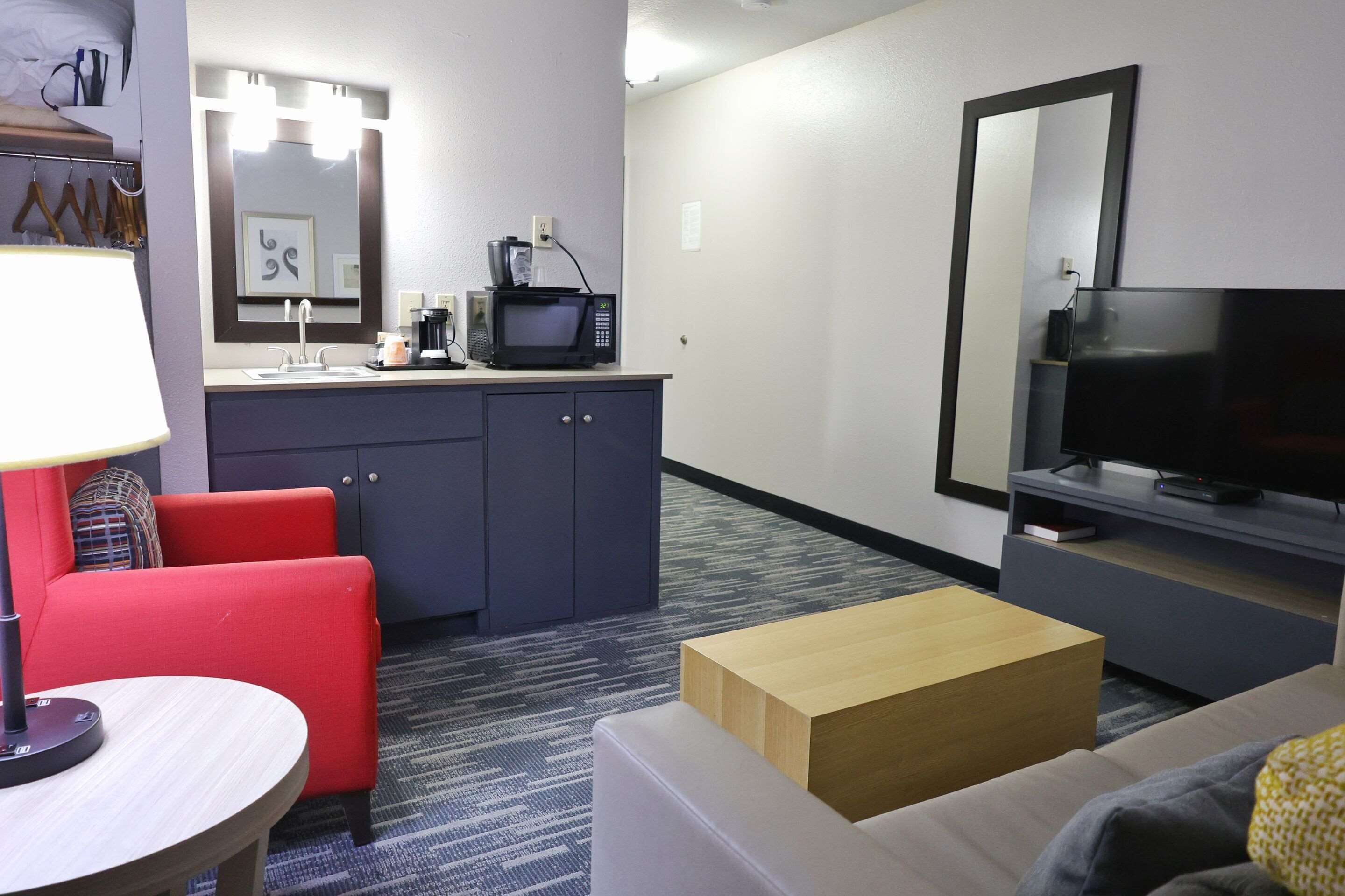 A hotel room with a kitchenette, a TV, a red armchair, a white sofa, and a lamp.