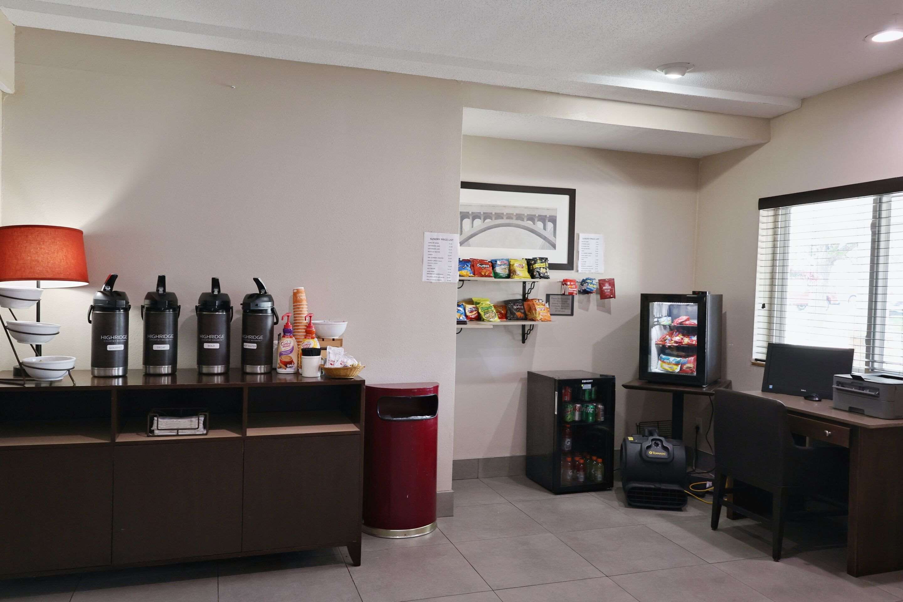 A hotel reception area with a counter, coffee makers, a trash bin, a refrigerator, a television, and a window with blinds.