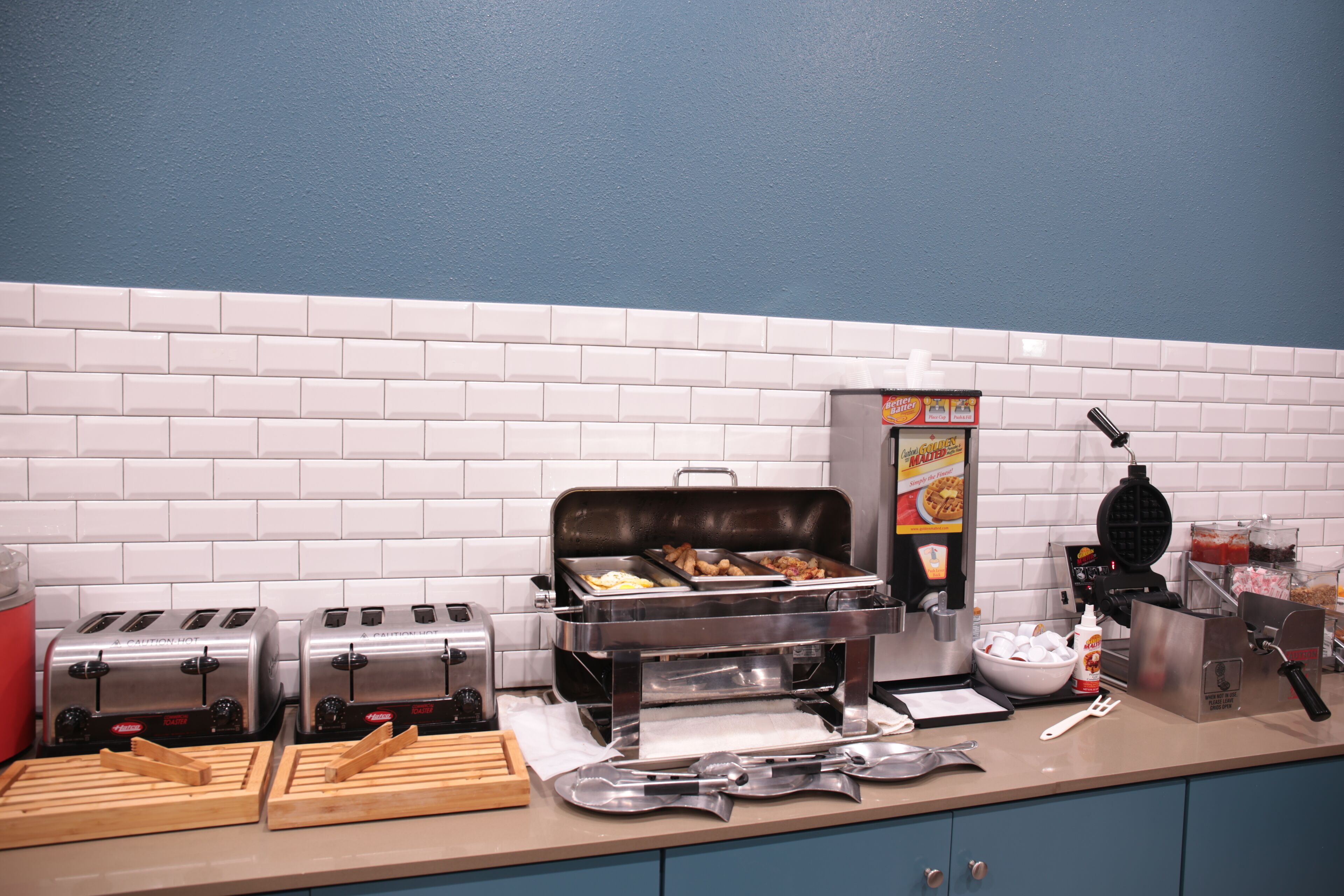 A kitchen with two toasters, a waffle maker, and a buffet station with trays of food.