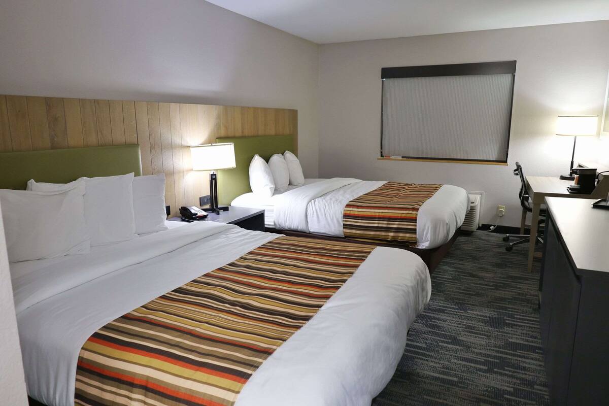 A hotel room with two beds, a desk, a chair, a lamp, and a large window with blinds.