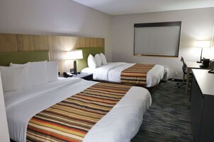 Habitación de hotel con dos camas, un escritorio, una silla, una lámpara y un ventanal grande con persianas.