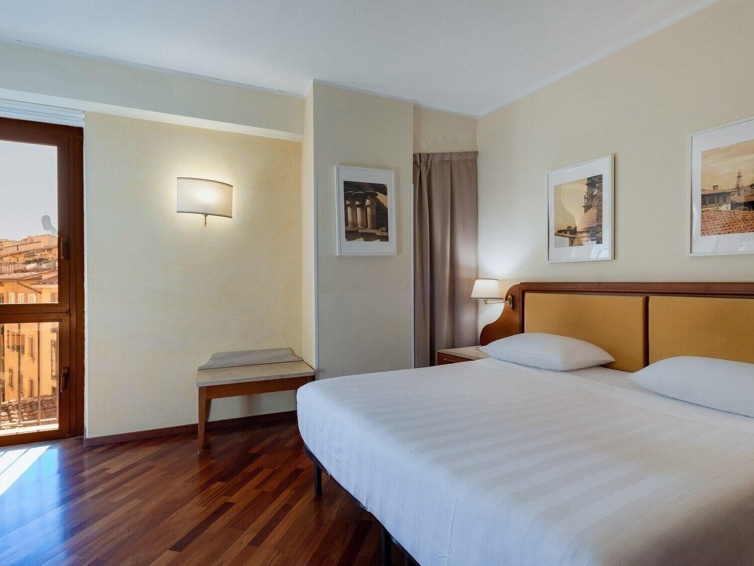Habitación de hotel con una cama grande, una mesita de noche, una lámpara, una ventana con vistas a edificios y cuadros en la pared.