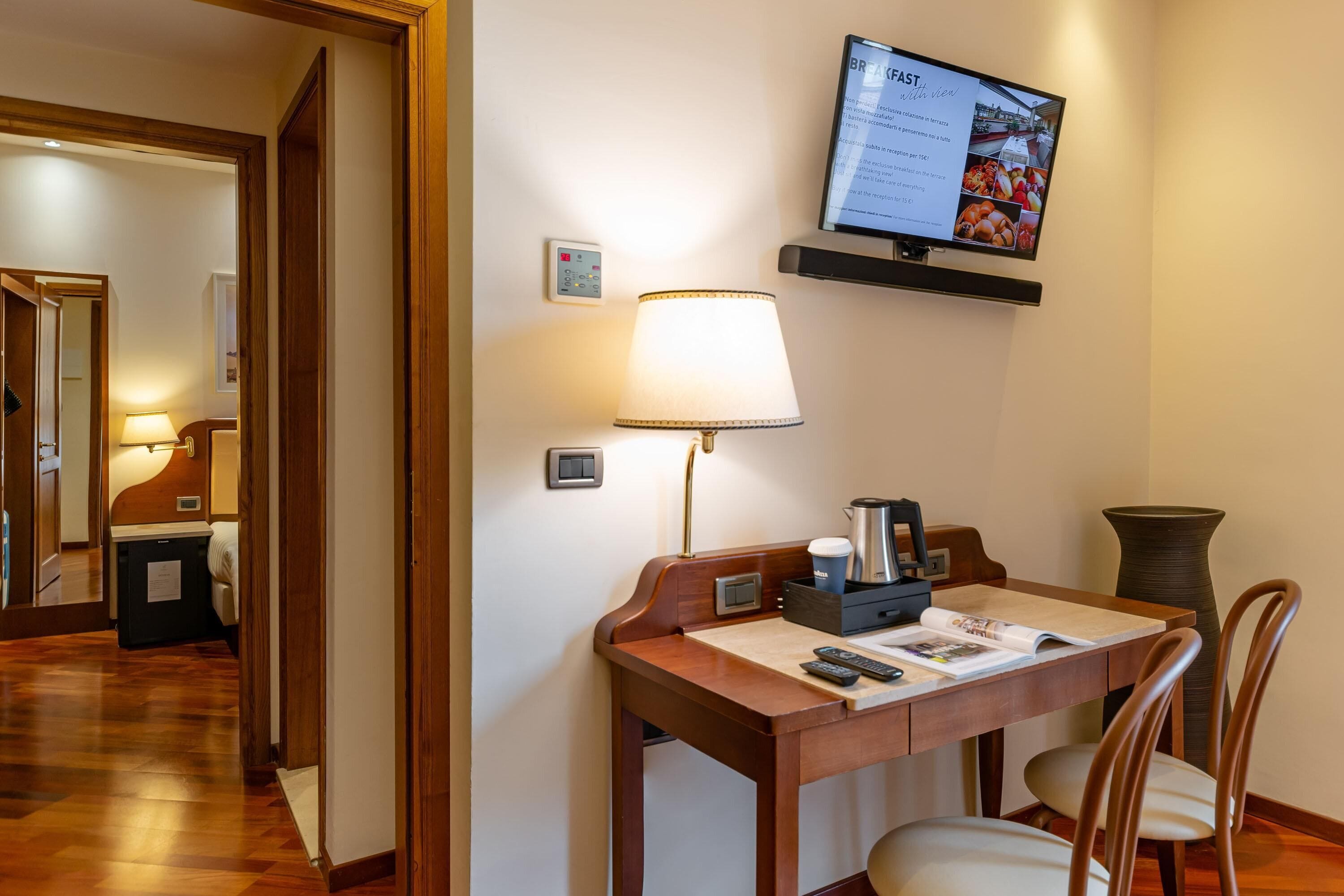 A hotel room with a wooden desk, a lamp, a television displaying a menu, a coffee maker, and a vase.