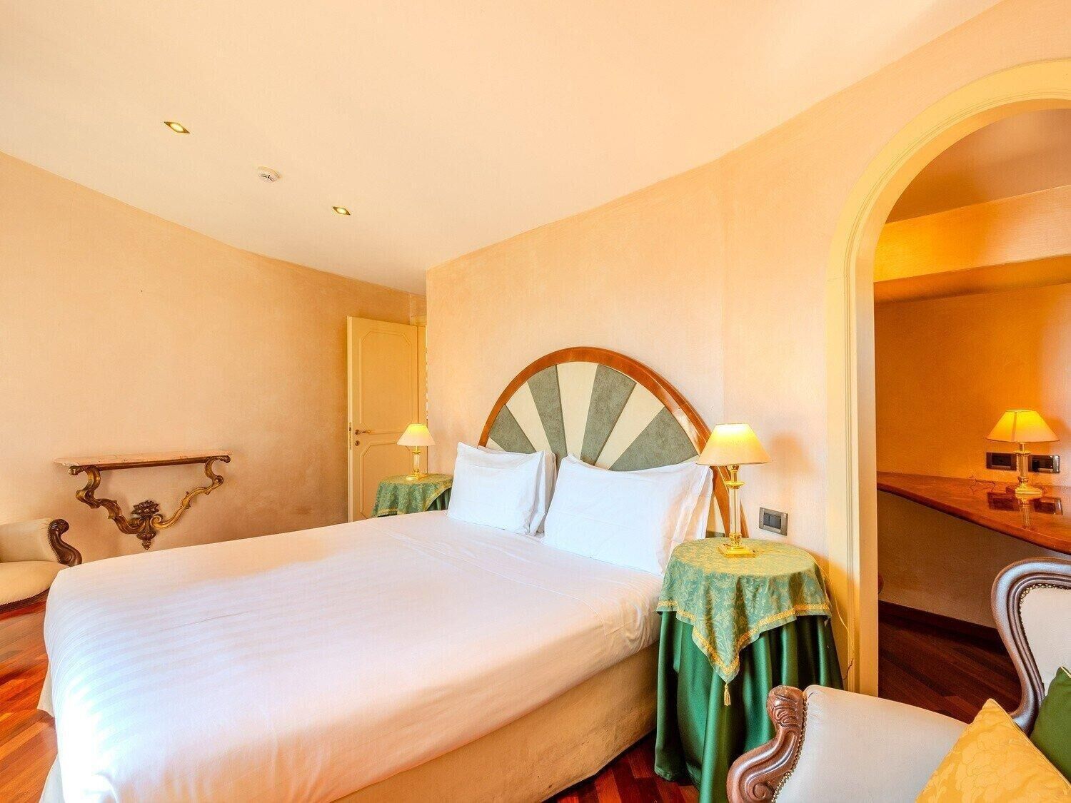 Una camera d'albergo con un letto grande, due comodini con lampade, una sedia di legno e uno specchio a parete.