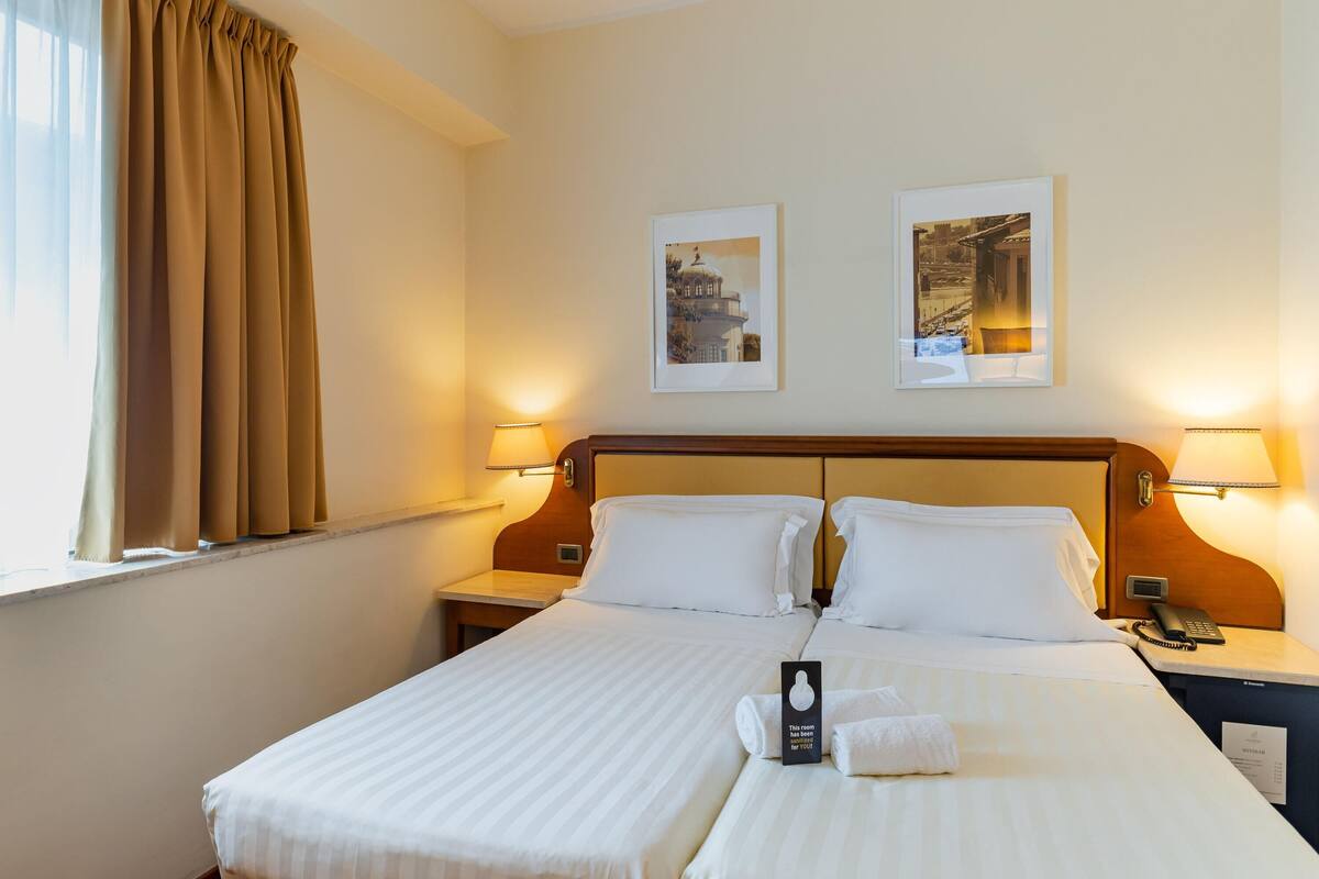 A hotel room with a double bed, two bedside tables, a nightstand, a telephone, a lamp, and two framed pictures on the wall.