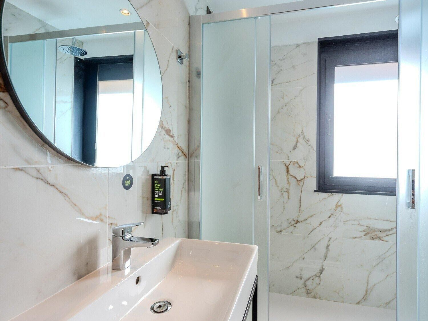 A modern bathroom with a marble sink, a round mirror, a handheld showerhead, and a glass-enclosed shower.