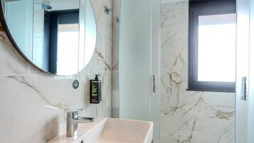 A modern bathroom with a marble sink, a round mirror, a handheld showerhead, and a glass-enclosed shower.