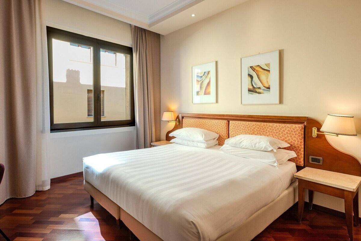 A hotel room with a large bed, two bedside tables, a window with curtains, and two framed artworks on the wall.