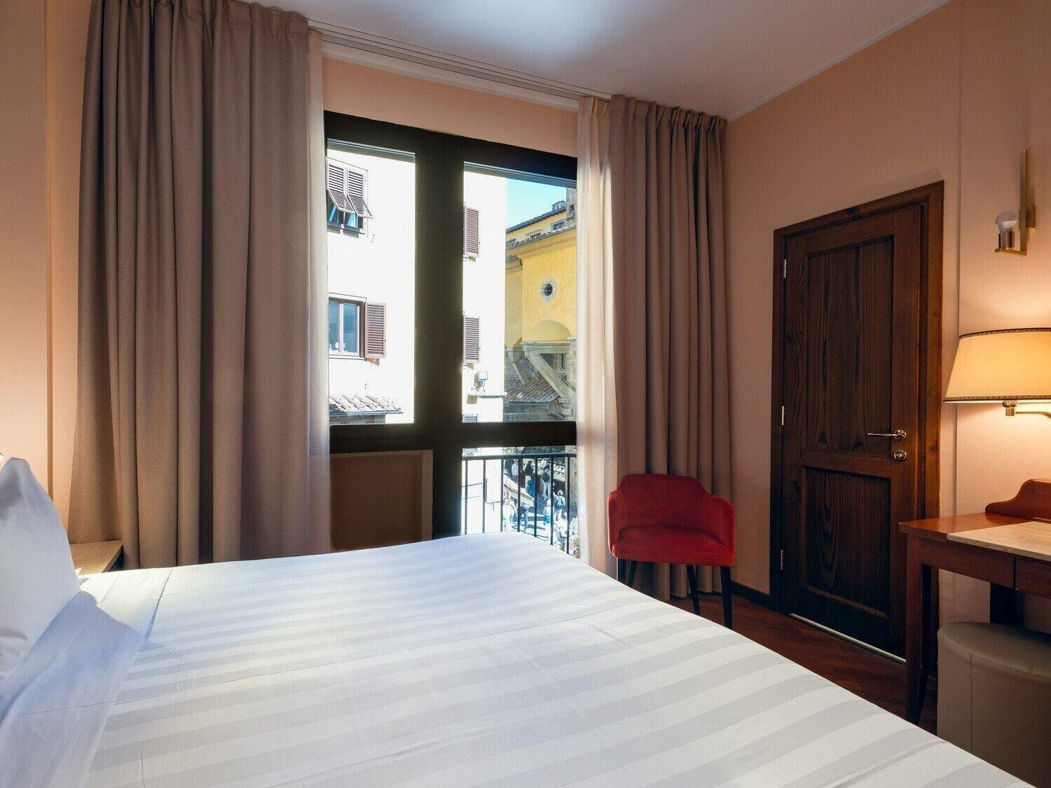 Une chambre d’hôtel avec un grand lit, un fauteuil rouge, un bureau en bois et une vue sur les immeubles par la fenêtre.