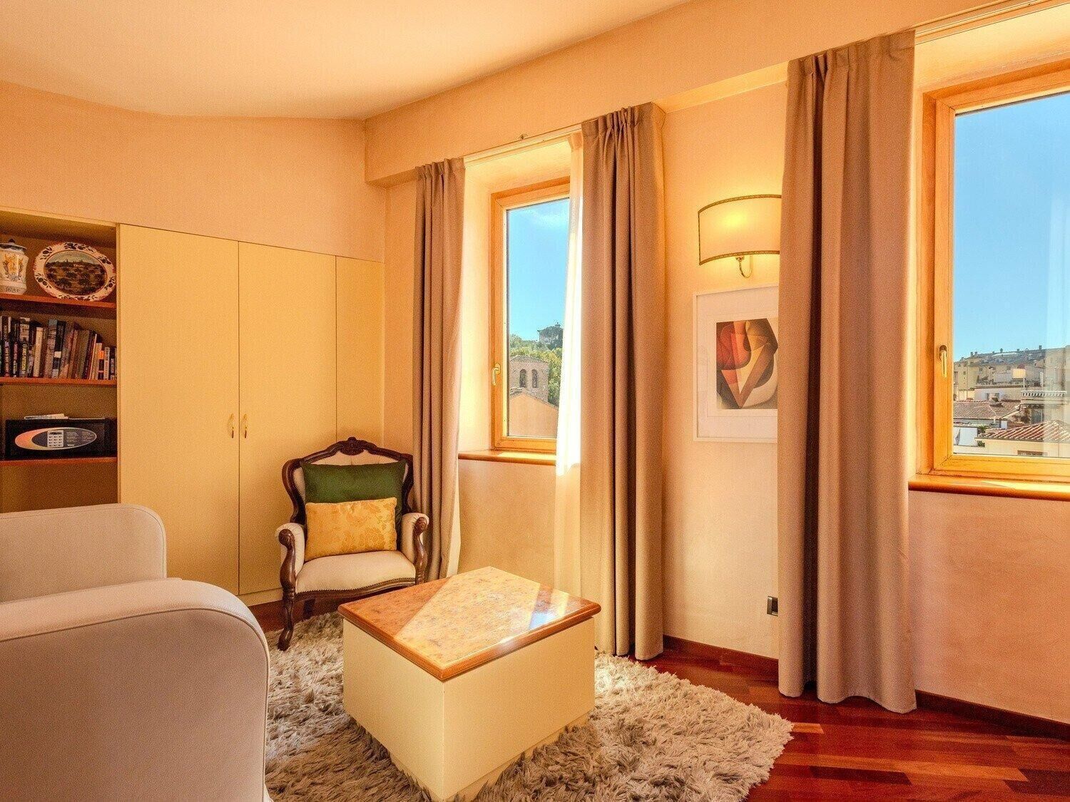 Ein gemütliches Wohnzimmer mit Sofa, Sessel und Hocker. Ein Bücherregal, ein Fenster mit Vorhängen und ein Gemälde an der Wand sind ebenfalls vorhanden.