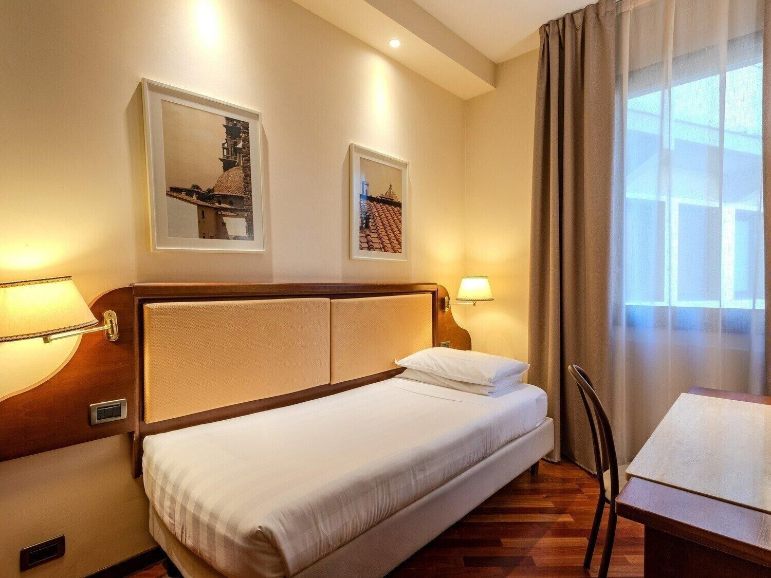 Habitación de hotel con una cama, dos lámparas fijadas a la pared, una mesa y dos cuadros enmarcados.