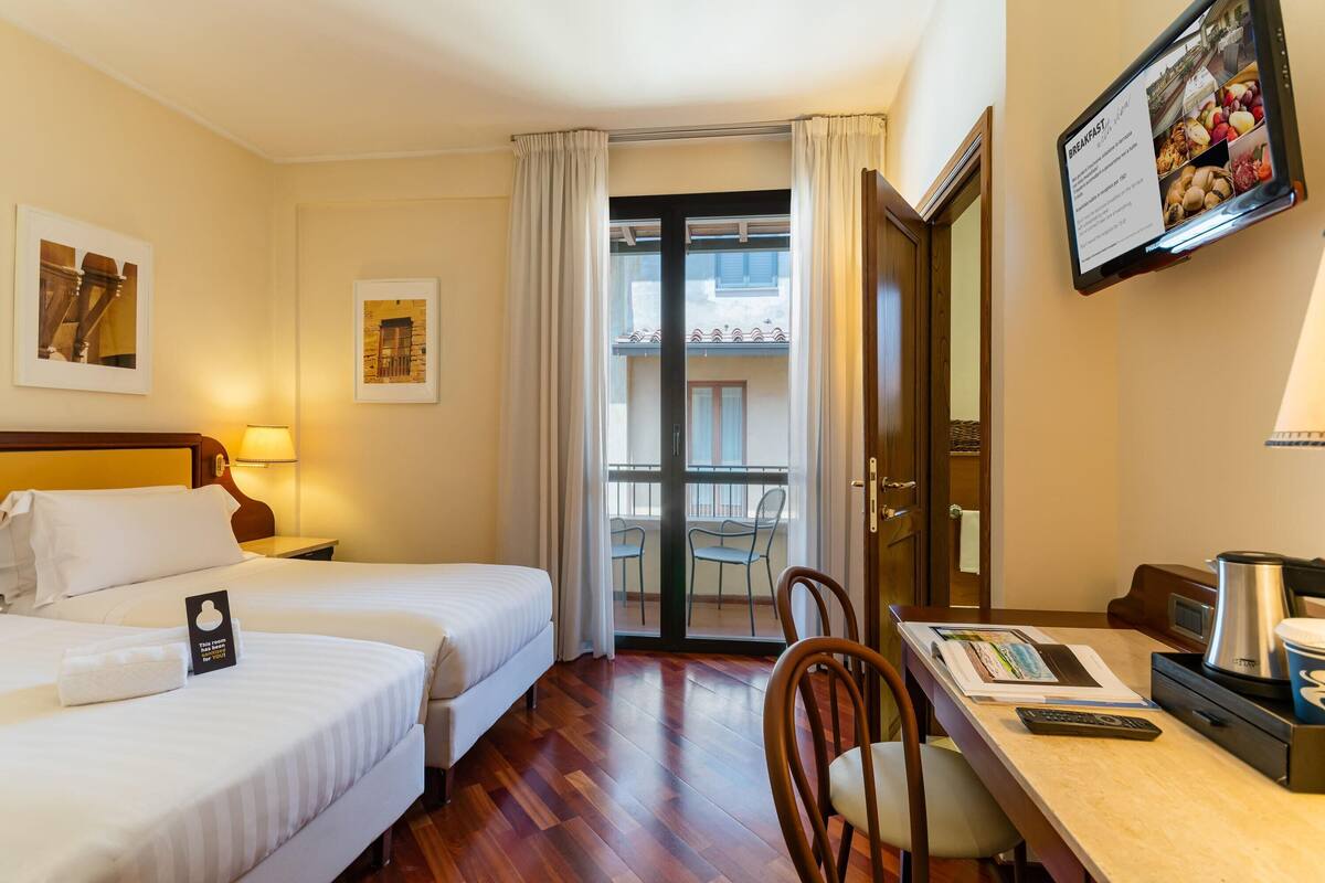 A hotel room with two beds, a desk with a coffee maker, a TV displaying a breakfast menu, and a balcony with chairs.
