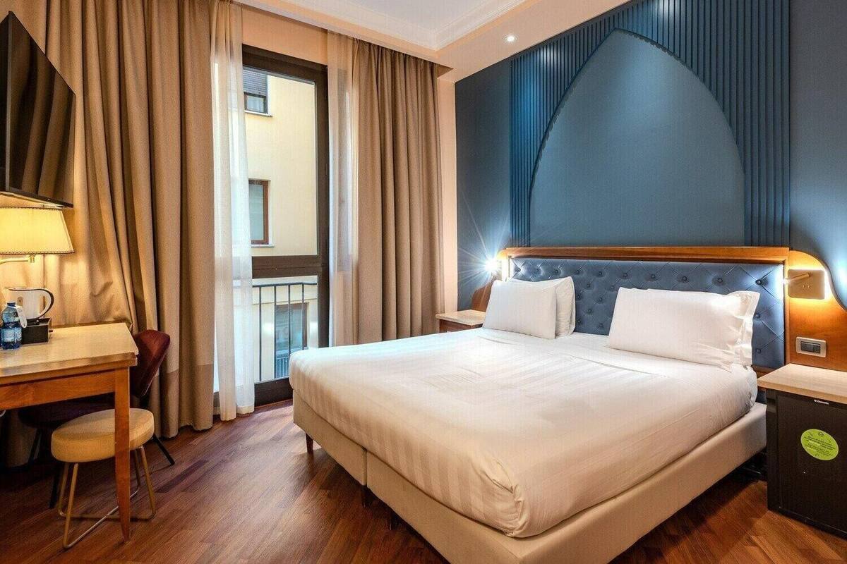A hotel room with a large bed, a desk with a lamp, and a view of a building through the window.