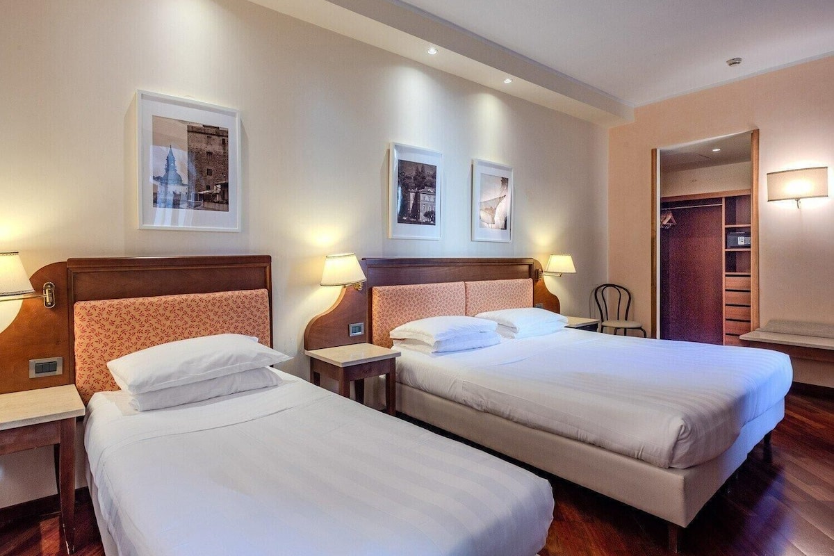 A hotel room with two single beds, a wooden headboard, bedside tables with lamps, and framed pictures on the wall.