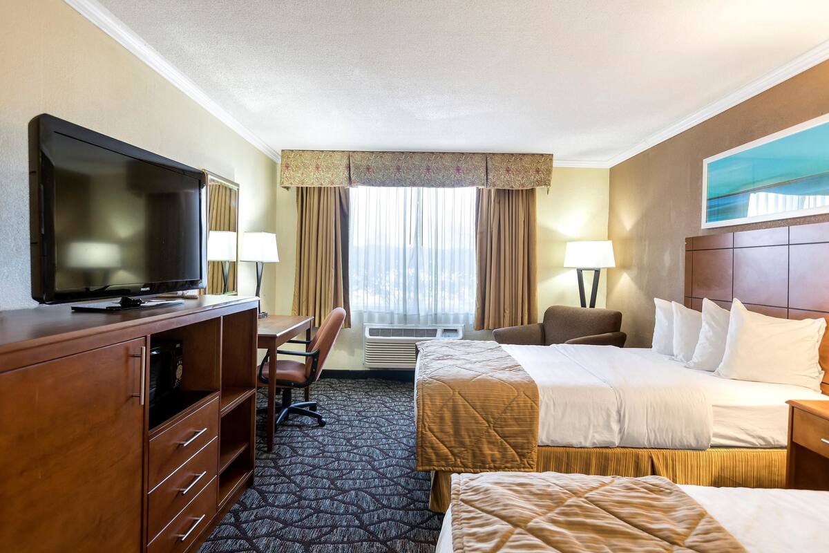 A hotel room with a TV, a desk, two beds, a chair, and a window with curtains.