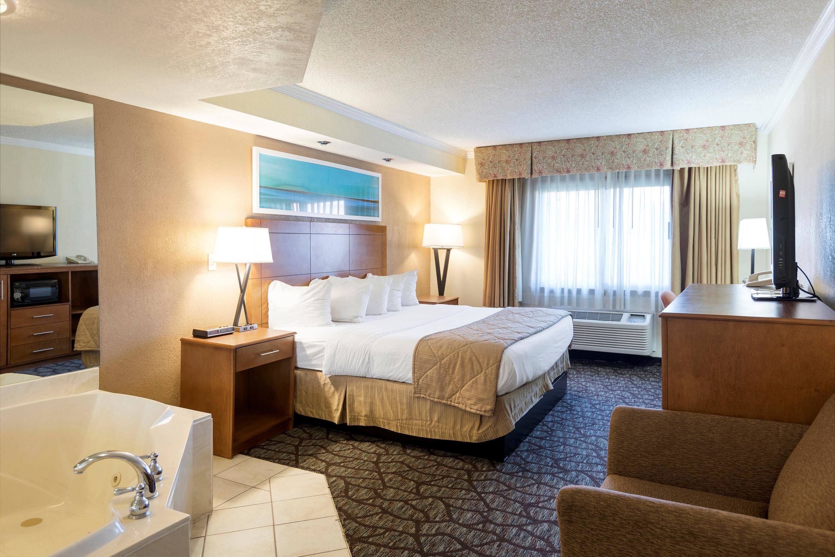 A hotel room with a large bed, two bedside lamps, a television, a desk, a chair, a bathtub, and a window with curtains.