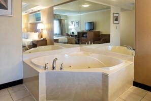 Un baño de hotel con una bañera grande, un espejo, una cama, un escritorio y un televisor.