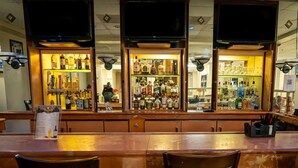 Una zona de bar con una barra alargada, varios estantes llenos de botellas de licor y un atril con la carta.