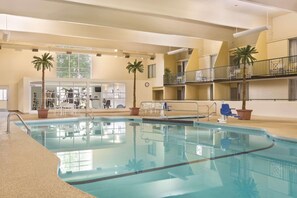 An indoor swimming pool with clear water, surrounded by a beige-colored interior, a seating area with tables and chairs, and two palm trees.