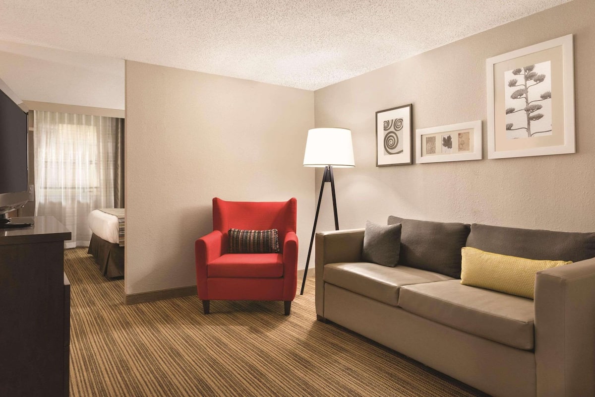 Una habitación de hotel con un sillón rojo, un sofá color beige, una lámpara de pie y cuadros en la pared.