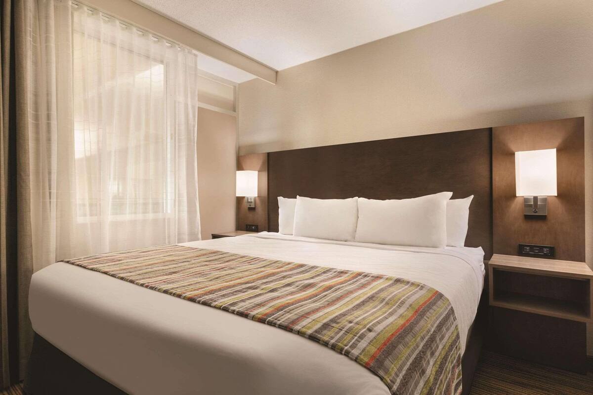 Habitación de hotel con una cama grande, dos mesitas de noche con lámparas, un cabecero y una ventana con cortinas traslúcidas.