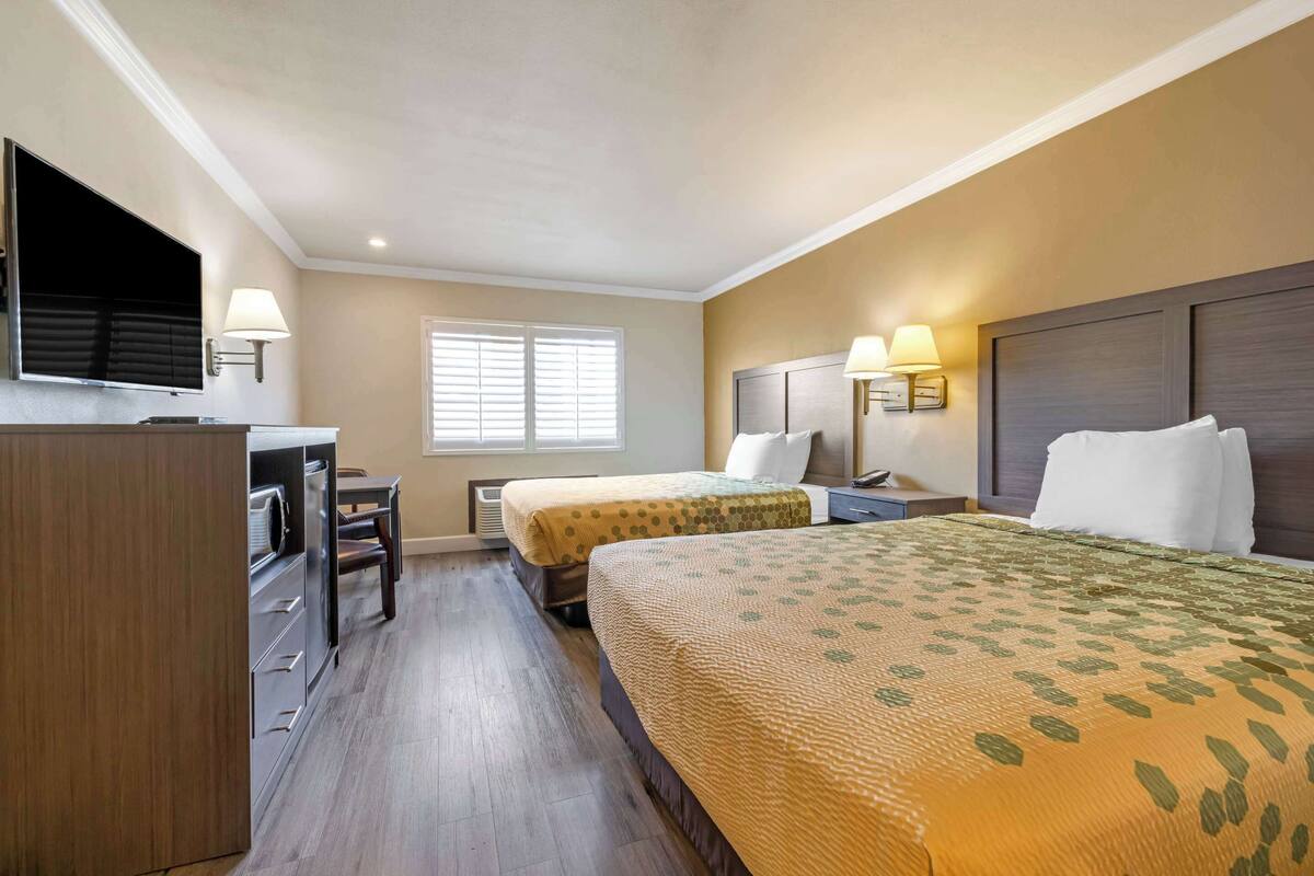 A hotel room with two beds, a desk, a TV, and a window with blinds.