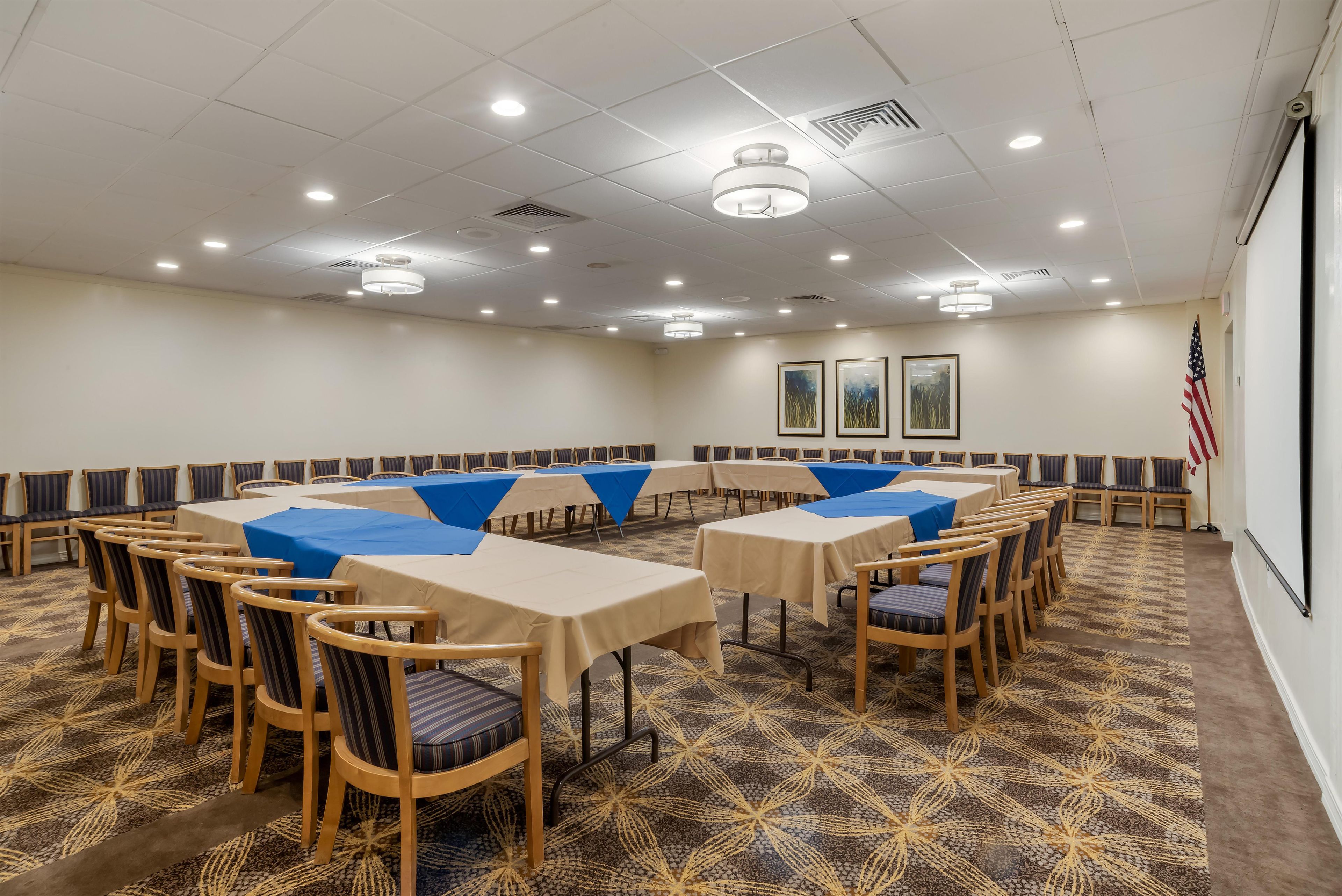 A conference room with multiple tables and chairs, arranged in a U-shape, with ceiling lights and an American flag on the wall.
