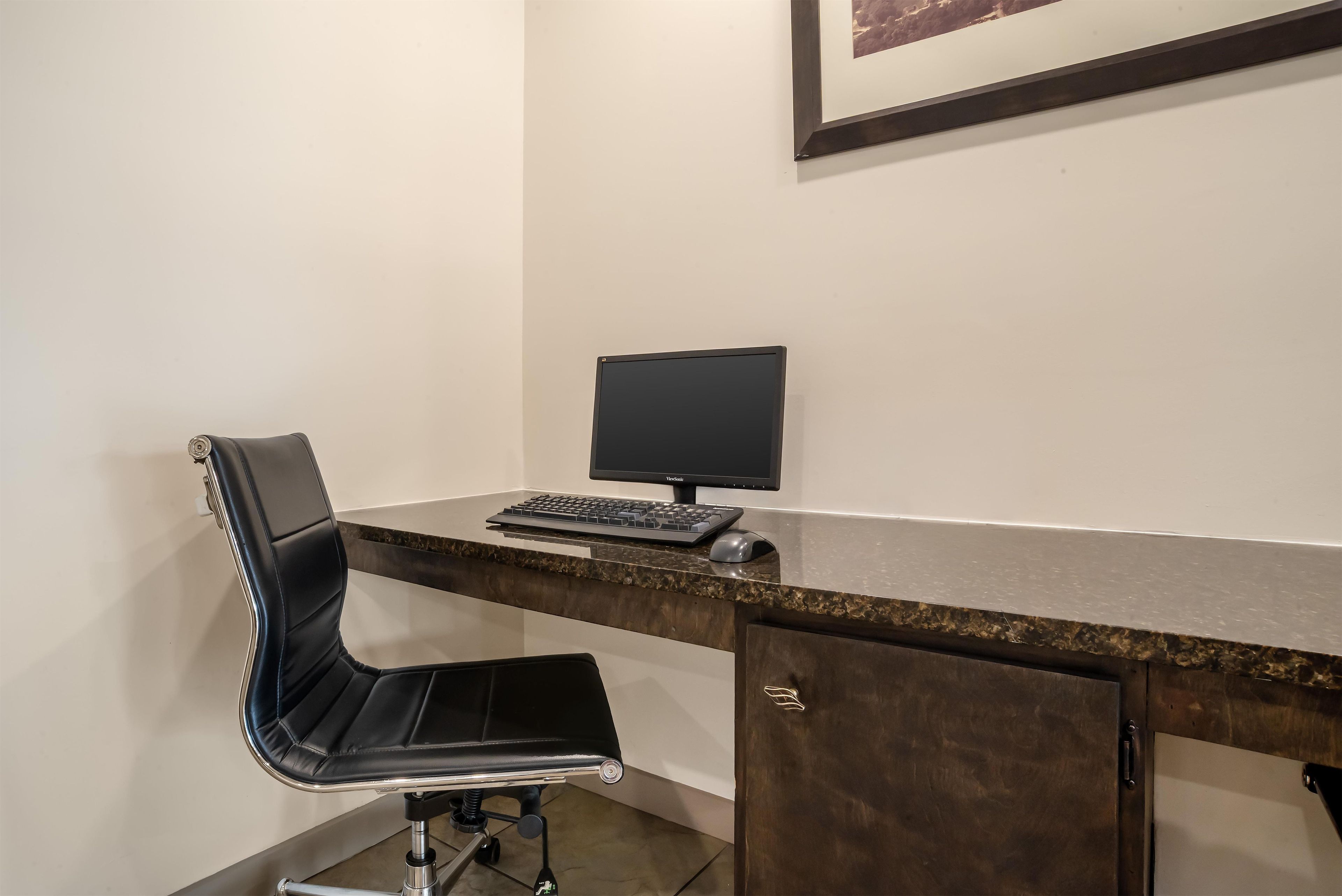 A corner office with a desk, a chair, a monitor, and a framed picture on the wall.