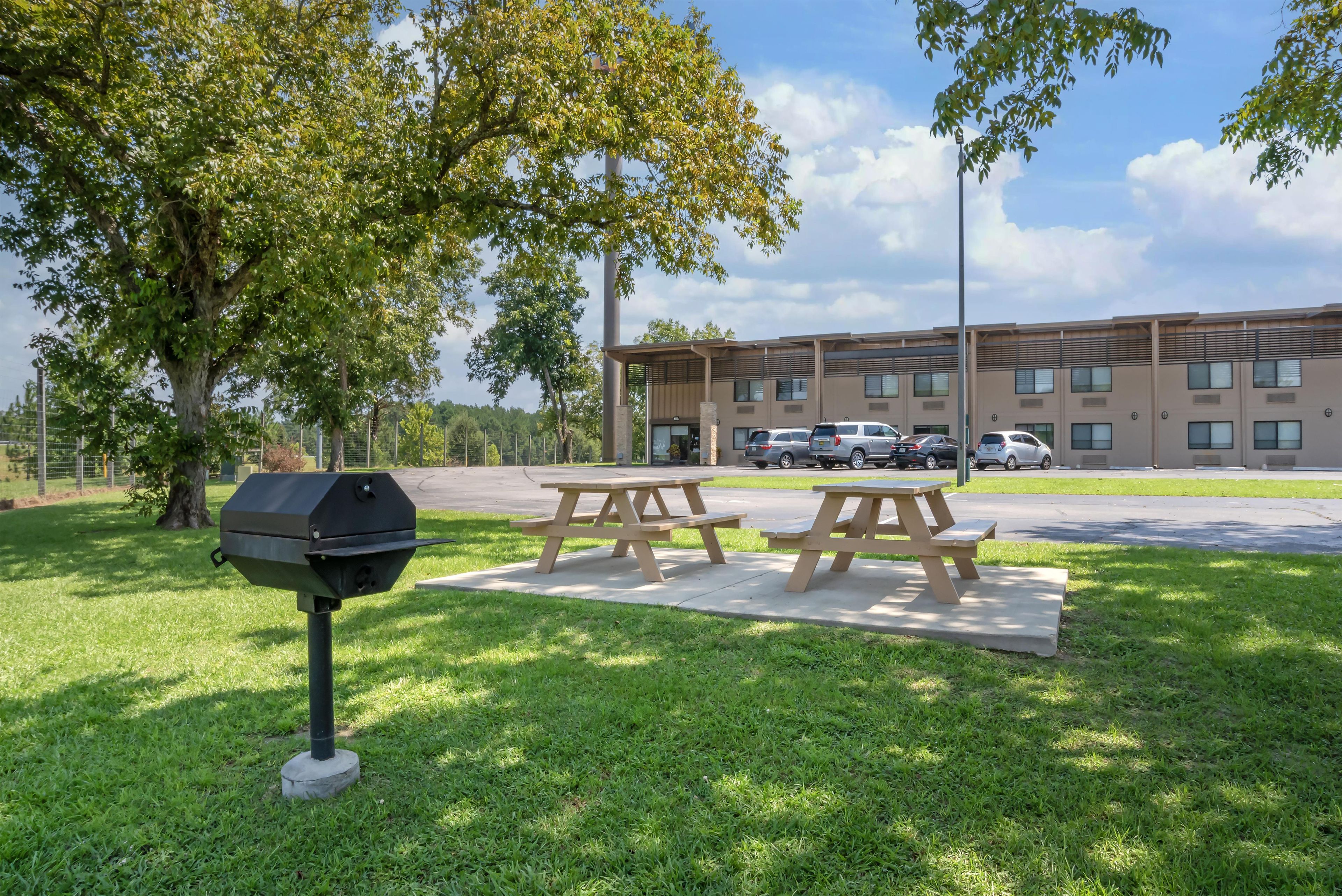 Un espace pique-nique équipé d’un barbecue et de tables, entouré de pelouse et d’arbres, avec un bâtiment visible au loin.