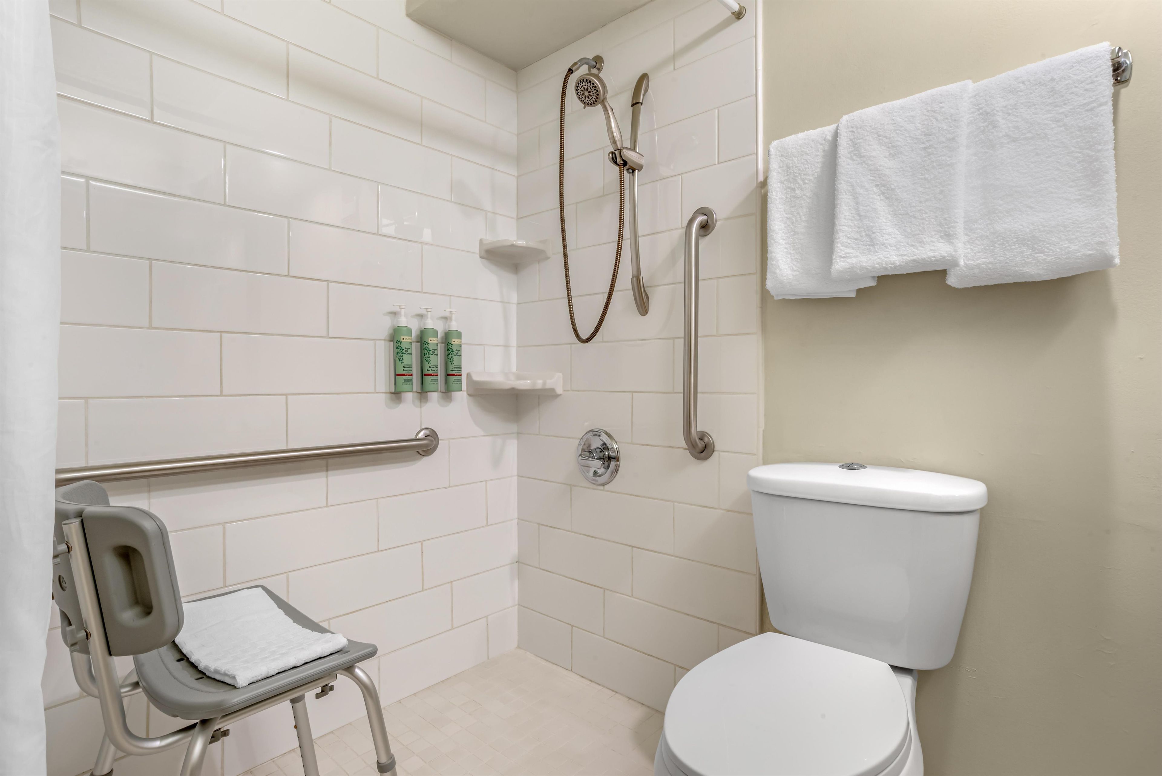 A bathroom with white tiles, a toilet, a shower with a handheld showerhead, a grab bar, and a small shelf with toiletries.