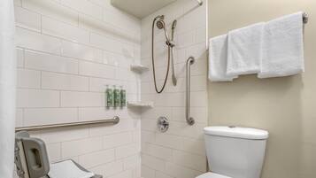 A bathroom with white tiles, a toilet, a shower with a handheld showerhead, a grab bar, and a small shelf with toiletries.