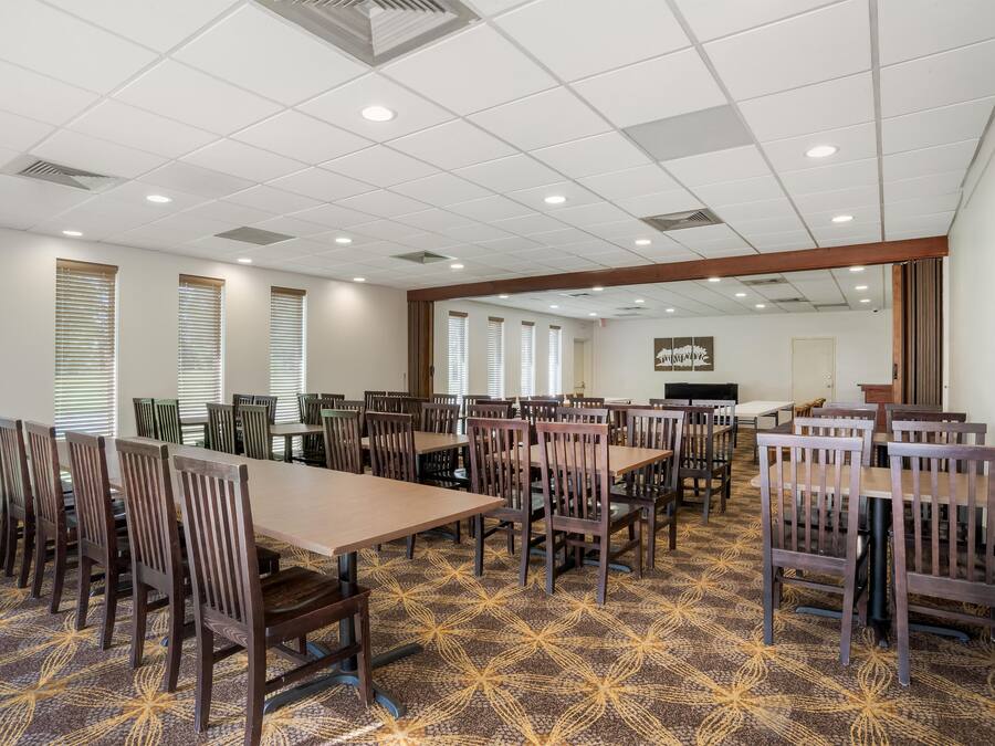 A conference room with multiple tables and chairs, large windows with blinds, and a patterned carpet.